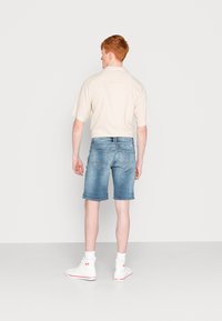 Chemise beige clair à manches courtes, shorts en denim bleu, et baskets montantes blanches. Texture en denim déchiré avec ourlet roulé sur les shorts.