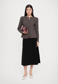 Brown herringbone blazer with a front zipper, paired with a black midi skirt and white pointed-toe flats, holding a burgundy clutch.
