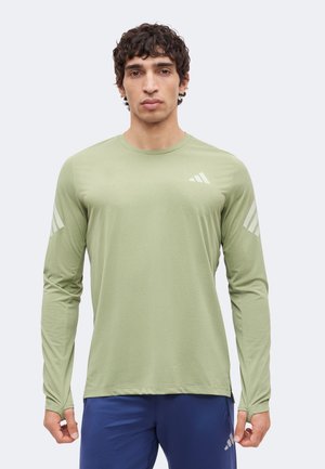 Tânăr cu păr creț purtând un tricou sportiv verde deschis cu mâneci lungi și pantaloni scurți albaștri pe un fundal simplu, deschis.