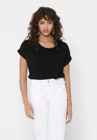 Top nero a maniche corte con scollatura rotonda, indossato da un modello con pantaloni bianchi. Il tessuto è morbido e il design è ampio.