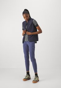 Marineblauwe fleece vest over een grijs T-shirt, gecombineerd met slanke paarse broek en groene wandelschoenen met oranje accenten. Casual buitenoutfit.