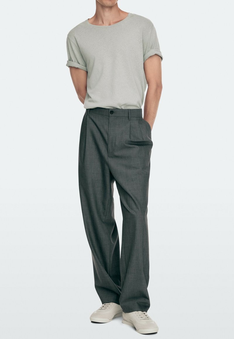 Man wearing light gray rolled-sleeve t-shirt, dark gray wide-leg trousers, and white sneakers, standing with hands in pockets.