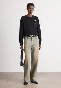 Sudadera negra con diseño de copa de cóctel, jeans de mezclilla clara de pierna ancha desgastados, bolso negro y zapatos negros de punta sobre un fondo neutro.