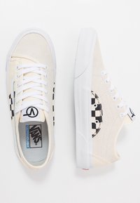 Cream canvas sneakers with white rubber soles. Features black checkerboard accents, a logo patch, and white laces. Textured surface.
