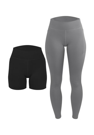 Frau steht barfuß und trägt ein schwarzes Crop-Top mit weißen Trägern sowie schwarze enganliegende Shorts vor einem einfarbigen hellen Hintergrund.