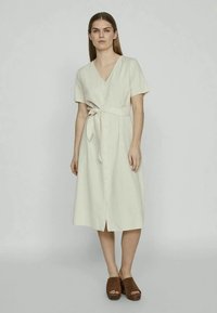 Γυναίκα που στέκεται φορώντας ένα μπεζ φόρεμα με κουμπιά μέχρι το γόνατο, κοντό μανίκι και ζώνη που δένει στη μέση, σε συνδυασμό με καφέ πλατφόρμες ανοιχτού δακτύλου.