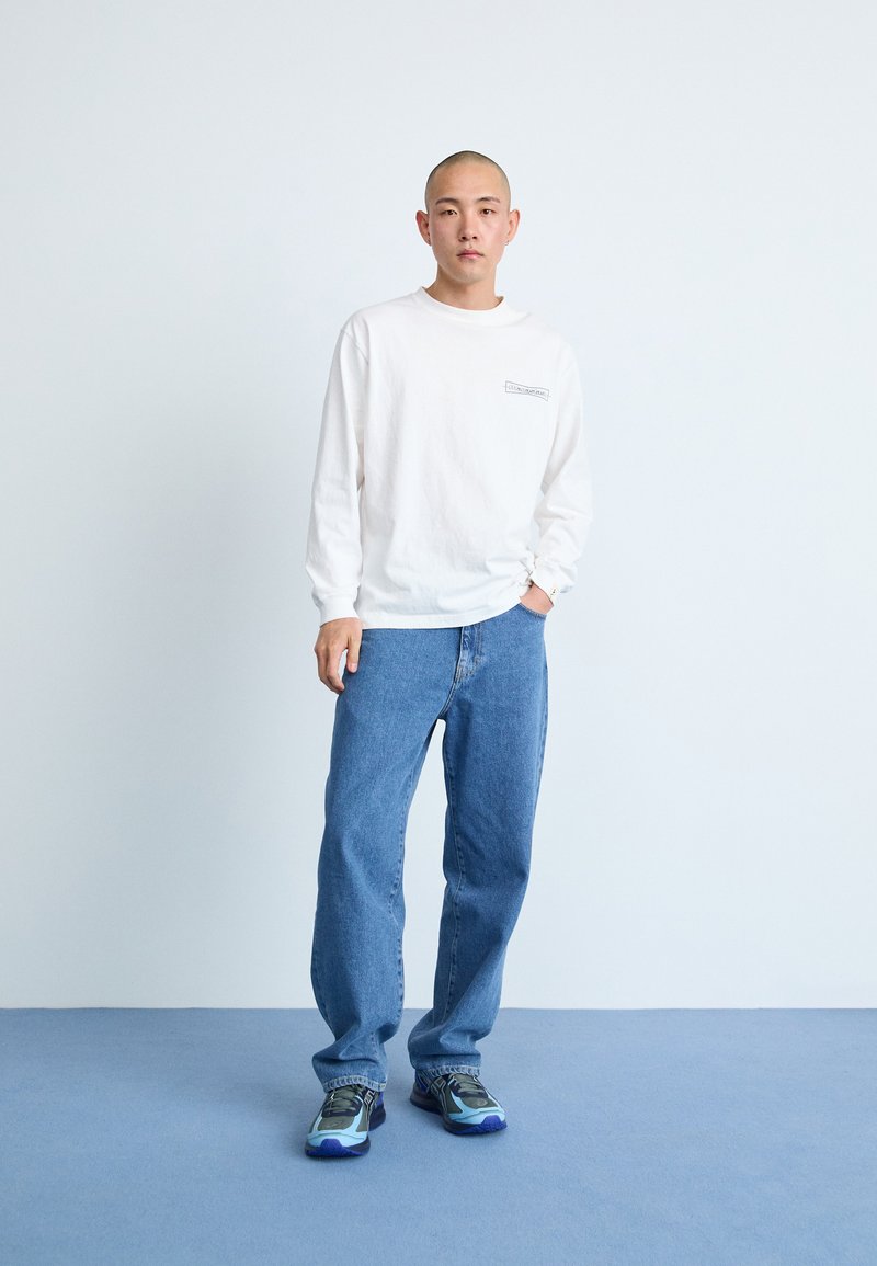 Chemise blanche à manches longues avec un petit logo, jeans en denim bleus et baskets multicolores ; debout sur un fond bleu clair.