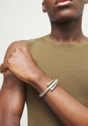 Persona con top sin mangas color oliva y pulsera ancha de plata en la muñeca izquierda, con los brazos cruzados sobre el pecho frente a un fondo neutro.