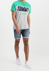 Camiseta de manga corta gris y verde con estampado "TOMMY JEANS NYC", combinada con pantalones cortos de denim enrollados y zapatillas blancas. Diseño sencillo y casual.