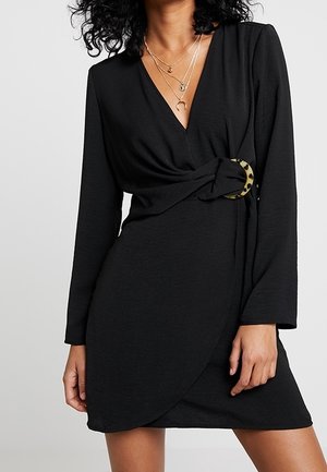 Femme portant une robe noire à manches longues en forme de portefeuille avec une boucle en écaille de tortue et des colliers en or superposés.