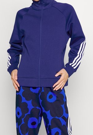 Person wearing a navy blue zip-up jacket with white stripes on sleeves and blue floral-patterned pants with black and blue flowers.