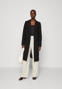 Lång svart blazer över en svart topp, parat med ljusa beige vida byxor. Modellen håller en krämfärgad väska med en slät yta.