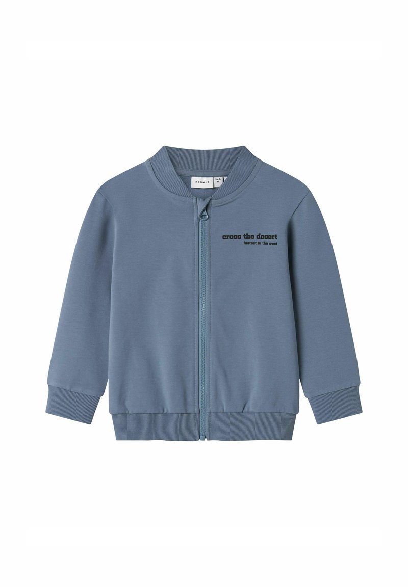 Zila jaku ar rāvējslēdzēju un ribotu apkakli un piedurknēm. Priekšpusē ir melns uzraksts "pār tuksnesi". Gluda auduma tekstūra.