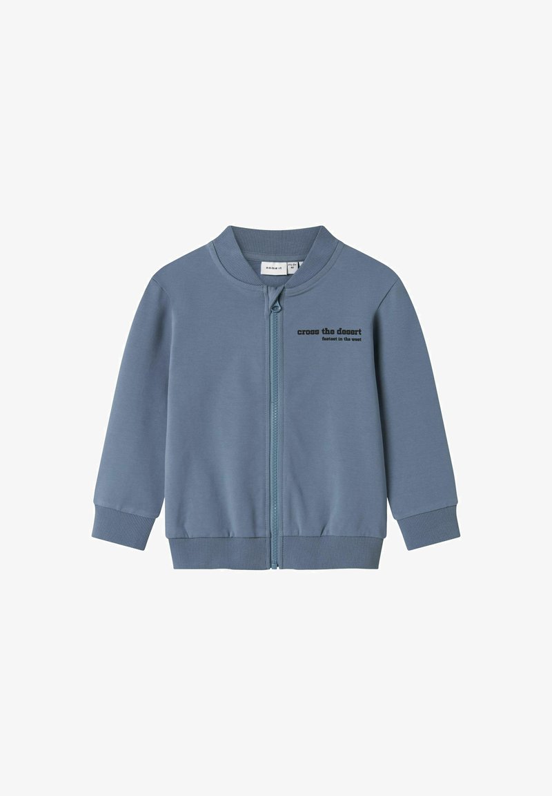 Zila jaku ar rāvējslēdzēju un ribotu apkakli un piedurknēm. Priekšpusē ir melns uzraksts "pār tuksnesi". Gluda auduma tekstūra.