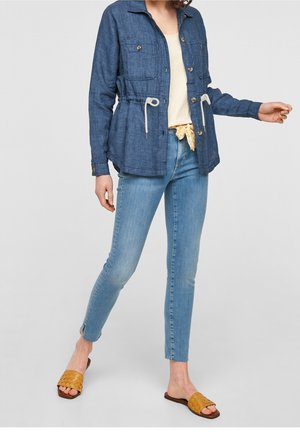 Femme portant une veste en jean bleue avec cordons, un jean skinny clair avec fentes aux chevilles, un top crème, des sandales tissées jaunes à enfiler.