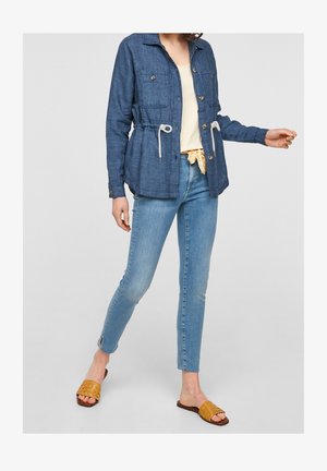 Mujer con chaqueta vaquera azul con cordones, vaqueros ajustados de lavado claro con aberturas en los tobillos, top crema, sandalias tipo slip-on tejidas amarillas.