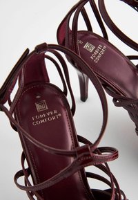 Burgundy high-heeled sandals with crisscross straps, smooth leather texture, and branded insoles. Features sleek pointed toes and slender heels.