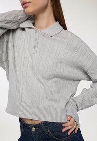 Kobieta z długimi włosami, ubrana w jasnoszary sweter z splotem i kołnierzykiem oraz ciemnoniebieskie dżinsy, pozująca z jednym ramieniem za głową.