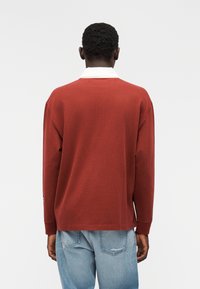 Μακρυμάνικη μπορντό πόλο μπλούζα με λευκό γιακά, με χαλαρή εφαρμογή και υφασμάτινη υφή, συνδυασμένη με ανοιχτό μπλε τζιν με σκισίματα.