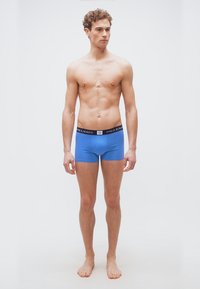 Νεαρός άνδρας όρθιος, ξυπόλητος, φορώντας μπλε boxer briefs Polo Ralph Lauren, μπροστά σε απλό λευκό φόντο.