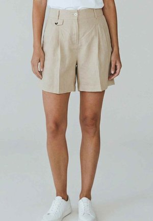 Persona che indossa pantaloncini beige a vita alta con pieghe e dettagli con bottoni, e sneakers bianche, in piedi davanti a uno sfondo chiaro e uniforme.