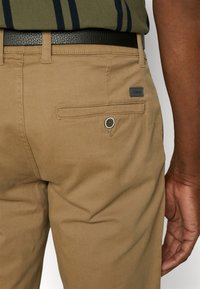 Gros plan d’une personne portant un pantalon beige avec une poche arrière boutonnée, une ceinture noire et une chemise rayée verte rentrée.