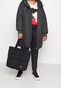 Person klädd i en svart vadderad kappa och svarta byxor, som håller en svart vadderad tygväska, stående mot en neutral vit bakgrund.