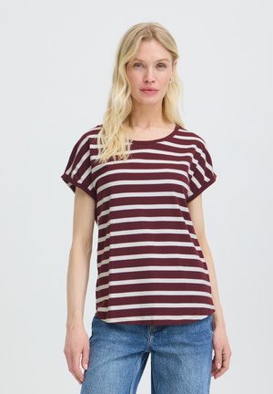 Blond kvinde iført en kortærmet skjorte med vinrøde og hvide striber og blå jeans, stående mod en ensfarvet lys baggrund.