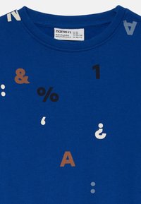 Camisa azul con símbolos impresos que incluyen un ampersand, un signo de porcentaje, el número uno, letras A y varios signos de puntuación en negro, blanco y marrón.