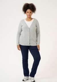 Grijze wol cardigan met knopen aan de voorkant, gecombineerd met een witte top en donkerblauwe jeans. Model draagt grijze en zwarte sneakers.