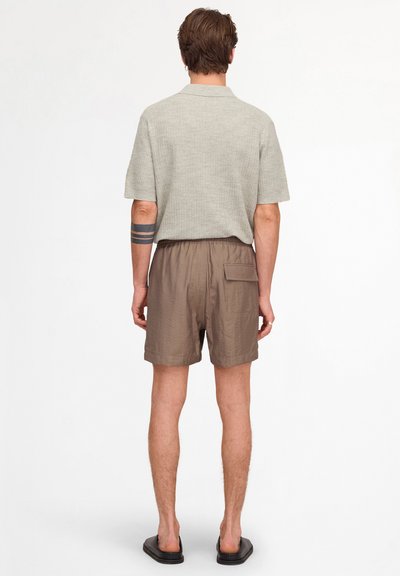 Homme de dos portant une chemise beige clair à manches courtes, un short marron avec une poche arrière, des chaussures noires sans lacets et un tatouage de bracelet noir sur le bras gauche.