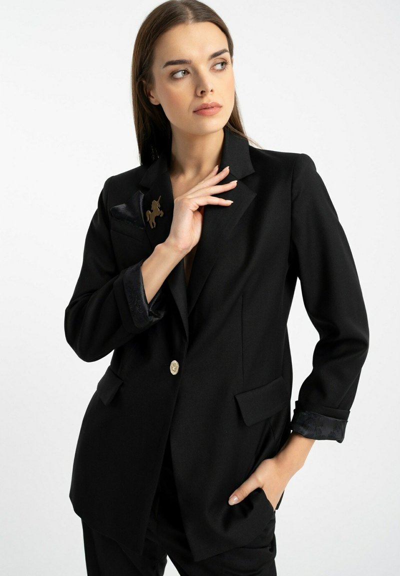Czarna marynarka o klasycznym fasonie, z klapami typu notch, jednolitym złotym guzikiem, dwiema przednimi kieszeniami oraz wzorzystymi mankietami z akcentem broszki.