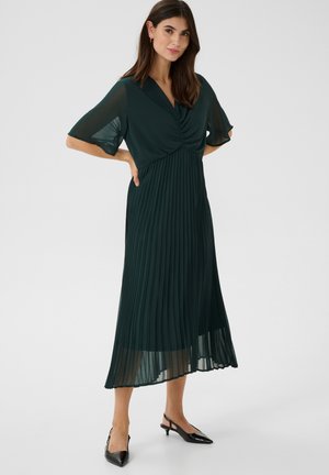 Pleated, mid-length dress in dark green fabric with a draped V-neck and flowing sleeves. Worn with black pointed-toe slingback heels.