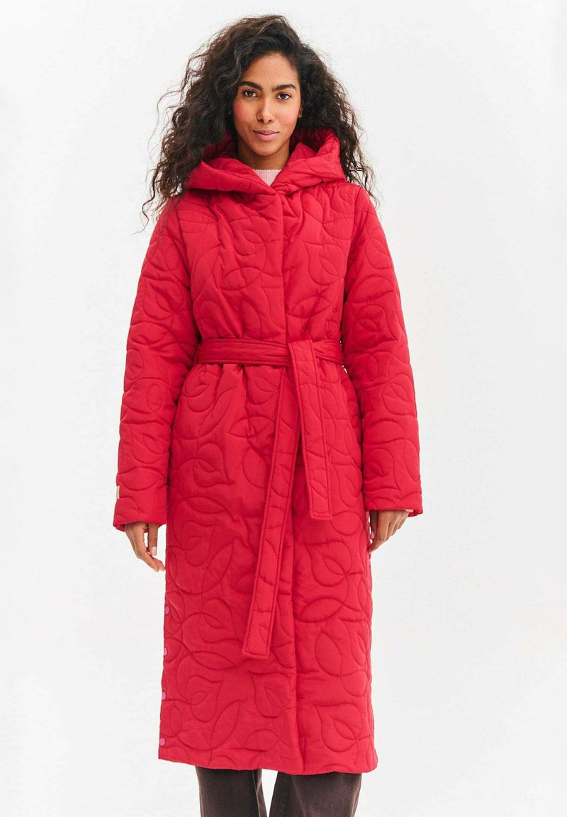 Czerwony pikowany płaszcz z kapturem, długimi rękawami i paskiem. Na tkaninie wytłoczony wzór liści. Z przodu guziki i kieszenie po bokach.