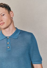 Blaues Strick-Poloshirt mit Kragen, drei Knöpfen zum Schließen und strukturierter Stoff. Kurze Ärmel mit geripptem Detail.