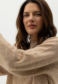 Világosbarna, puha kötött pulóver laza szabással és hosszú ujjakkal. A textúra bolyhosnak tűnik, ami kellemetlenséget és melegséget ad a dizájnnak.