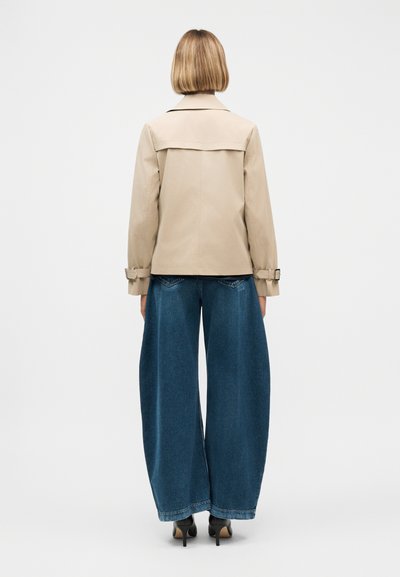 Femme aux cheveux blonds courts portant une veste beige, un jean large bleu et des chaussures à talons noires, debout de dos sur fond blanc.