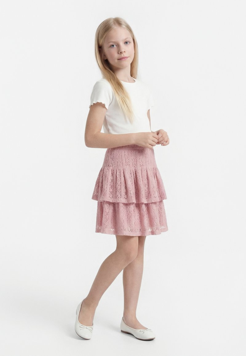 Jeune fille debout, portant un haut blanc à manches courtes, une jupe en dentelle rose à volants et des ballerines blanches, sur fond blanc.