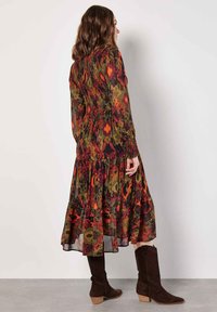 Langes, fließendes Kleid mit einem mehrfarbigen Blumenmuster in Rot-, Grün- und Gelbtönen. Verfügt über ein gerafftes Oberteil und lange Ärmel. Braune Wildlederstiefel.