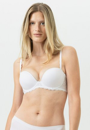 Blondine draagt een wit gewatteerd beha met kanten details, poserend tegen een effen lichte achtergrond.