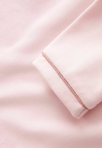 Tissu rose clair avec un bord plié, détaillé par une seule couture fine et brillante de fil rose le long du pli.