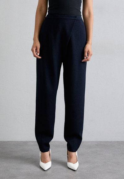 Pantalons taillés bleu marine avec une taille haute et une coupe décontractée, confectionnés dans un tissu doux. Associés à des talons aiguilles blancs, mettant en valeur la cheville.