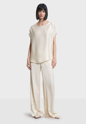 Femme aux cheveux noirs coupe au carré, portant un haut ample en satin couleur crème et un pantalon large, debout devant un fond clair uni.