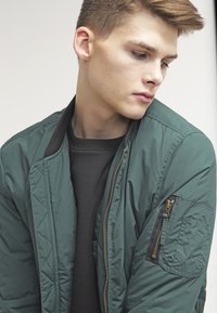 Grøn bomberjakke med lynlås foran, diagonalt quiltet mønster, ribbet krave og lynlåslomme på ærmet. Båret over en sort sweatshirt.