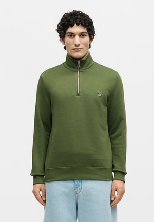 Man wearing olive green quarter-zip pullover with small embroidered logo on chest, paired with light blue jeans, standing against white background.