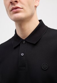 Juodas polo marškinėliai, pagaminti iš tekstūruoto audinio, su apykakle, dviem sagomis ir mažu apvaliu logotipu kairėje pusėje.