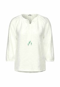 Blouse en lin blanc avec des manches longues, un col froncé et des liens ajustables présentant des accents verts. Coupe légèrement ample avec un ourlet arrondi.