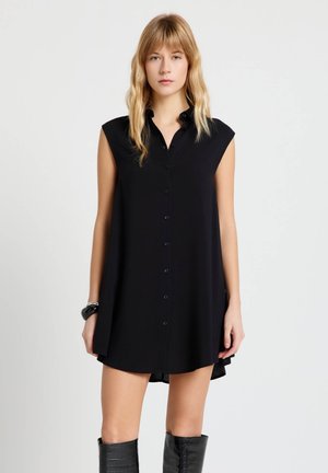 Femme aux cheveux blonds portant une robe noire sans manches boutonnée sur le devant et des bottes noires montantes jusqu'aux genoux, debout devant un fond blanc uni.