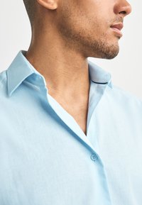 Nahaufnahme des Kieferbereichs und Nackens eines Mannes, der ein hellblaues Hemd mit offenem oberstem Knopf trägt, vor weißem Hintergrund.