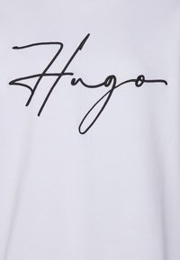 T-shirt bianco con un logo "Hugo" in corsivo nero. Il materiale è liscio con una trama semplice e il design è minimalista.